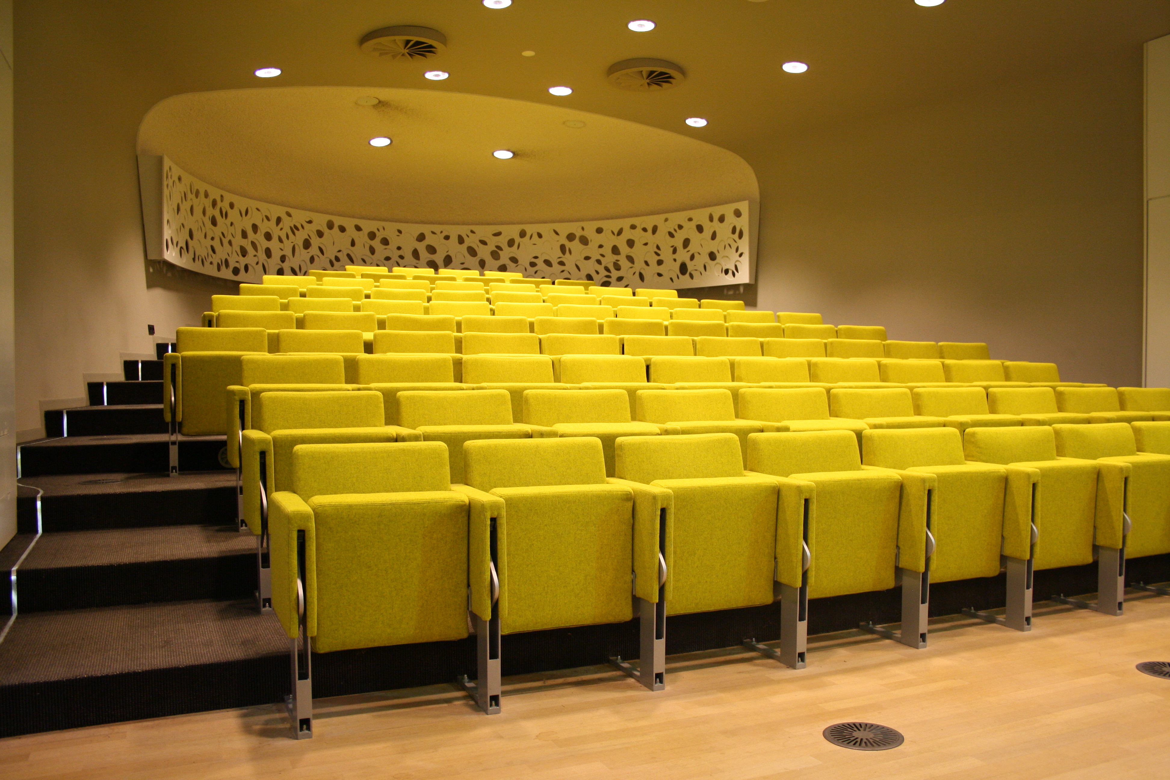 Op de foto is een congreszaal te zien, vol met gele stoelen. 