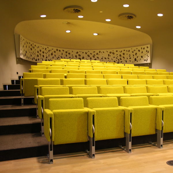 Op de foto is een congreszaal te zien, vol met gele stoelen. Op de foto is een congreszaal te zien, vol met gele stoelen.
