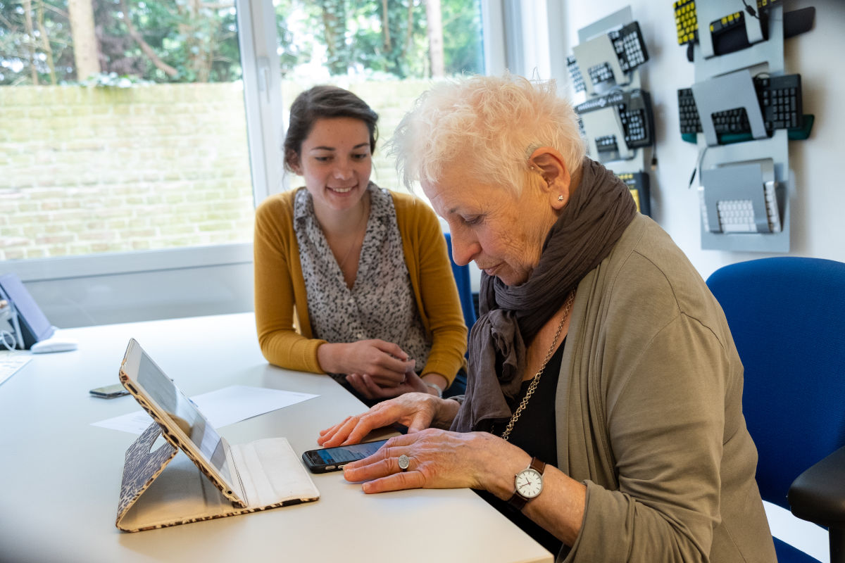 Oudere mevrouw krijgt ondersteuning in het gebruiken van haar telefoon en iPad. 