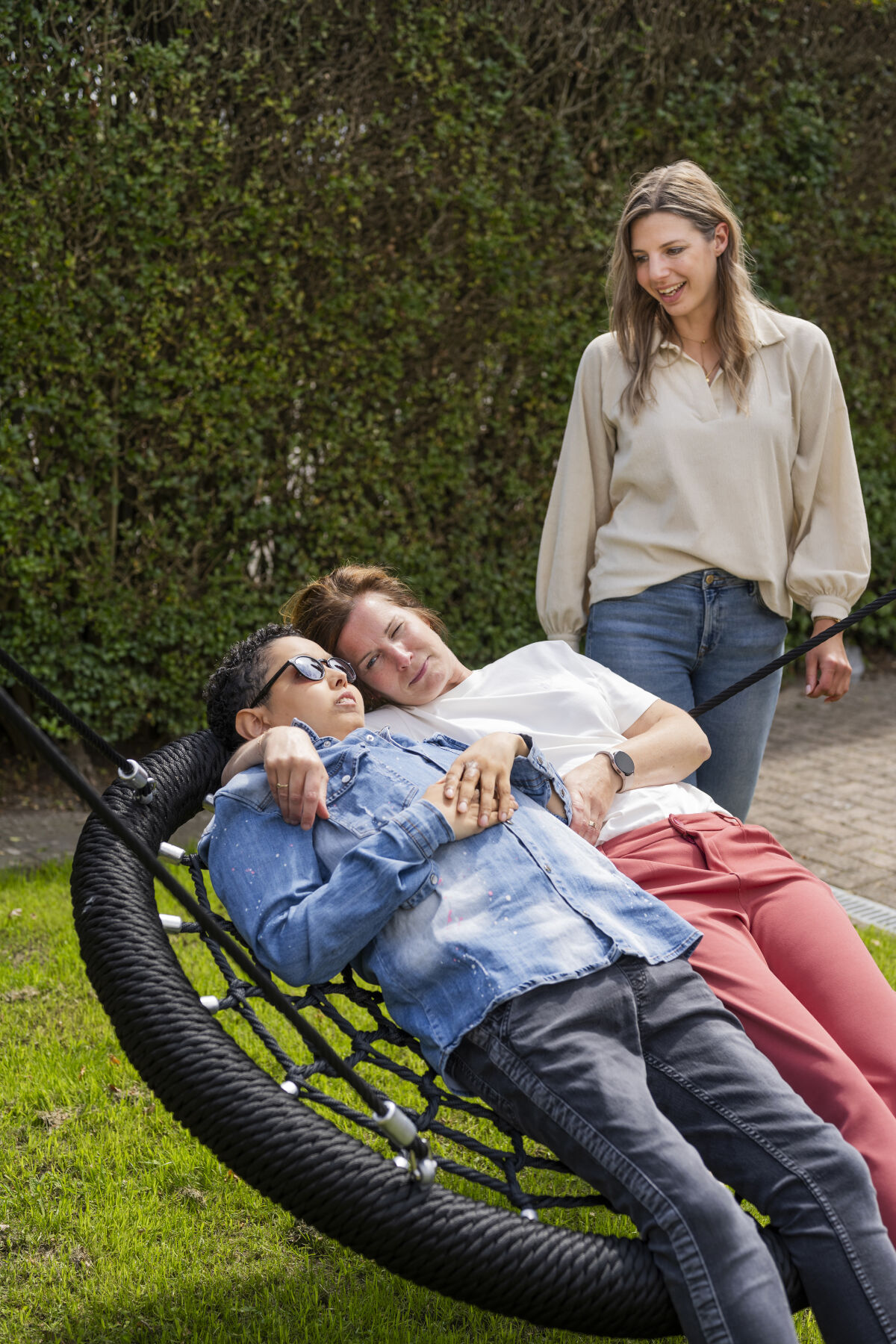 Cliënt van Visio ligt in een mandschommel en schommelt samen met een medewerker, op de achtergrond kijkt iemand glimlachend toe