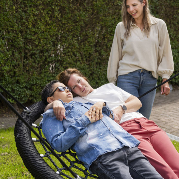 Cliënt van Visio ligt in een mandschommel en schommelt samen met een medewerker, op de achtergrond kijkt iemand glimlachend toe Cliënt van Visio ligt in een mandschommel en schommelt samen met een medewerker, op de achtergrond kijkt iemand glimlachend toe