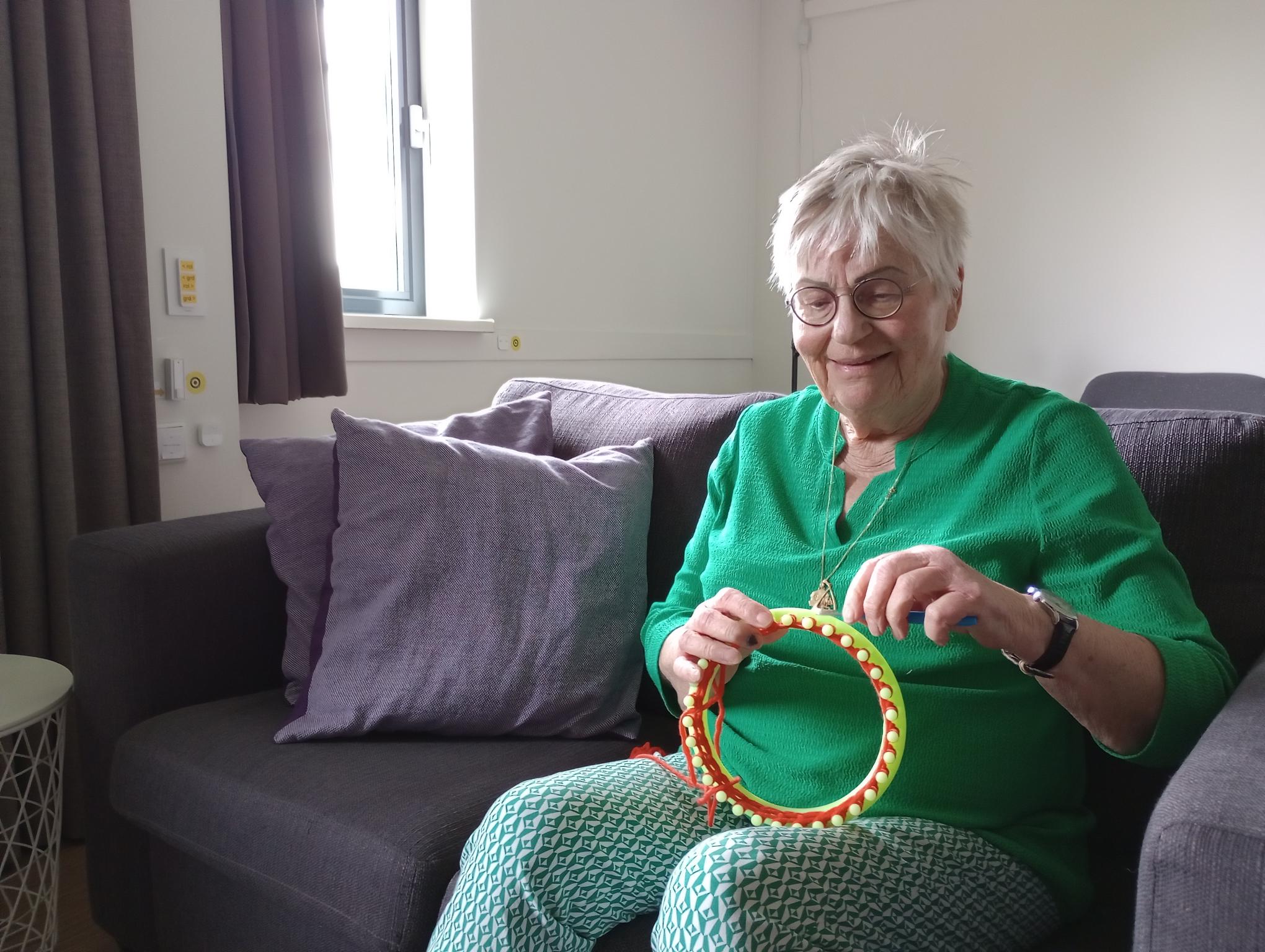 Foto van Sannie die met een breiring creatief aan de slag is, zij draagt een broek met een groen-wit patroon, een groene blouse met mouwen en een bril.