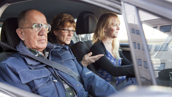 Drie personen zitten in een auto, de bijrijder draagt een bioptisch telescoopsysteem Drie personen zitten in een auto, de bijrijder draagt een bioptisch telescoopsysteem