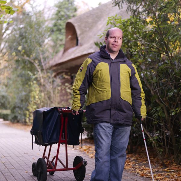 Cliënt brengt de post rond met een taststok in zijn hand en aan zijn andere hand een postwagentje Cliënt brengt de post rond met een taststok in zijn hand en aan zijn andere hand een postwagentje