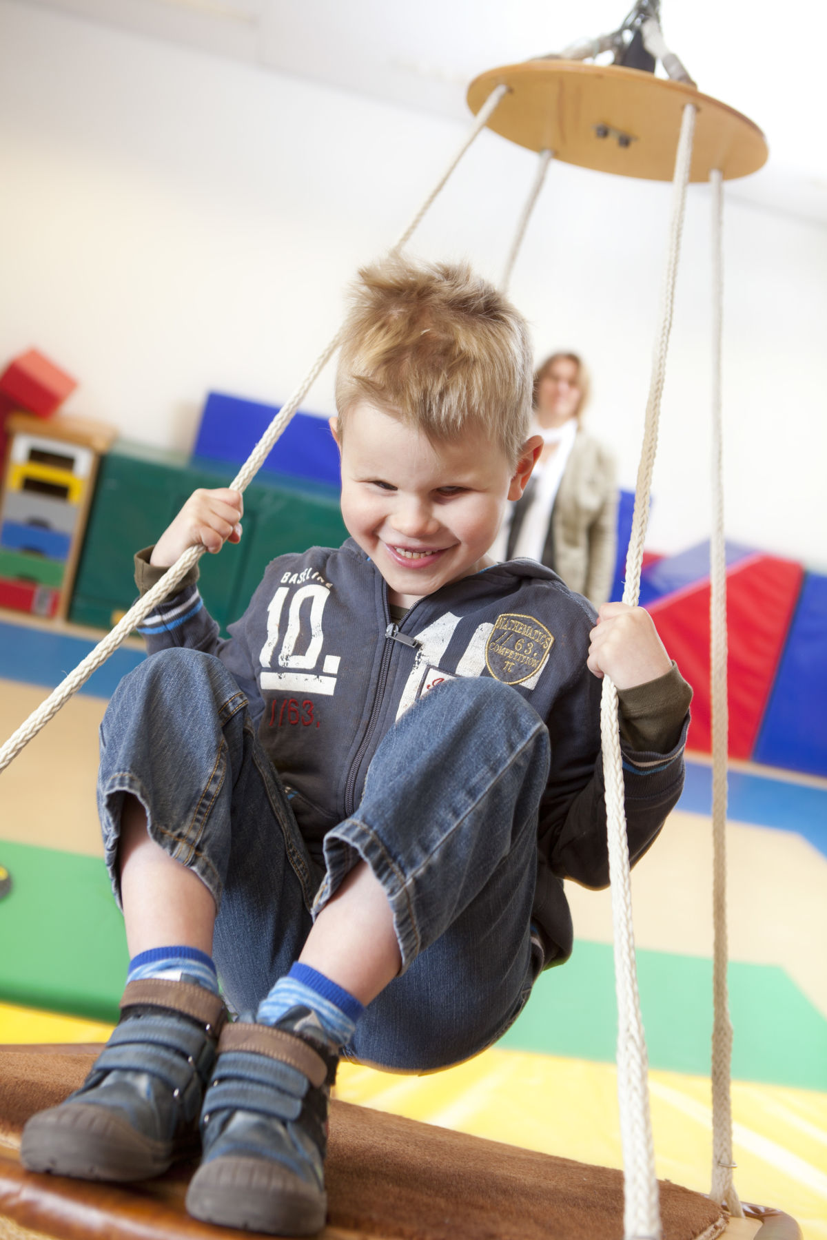 Een jongetje zit op een schommel. In de achtergrond zijn kleurrijke matten te zien. 