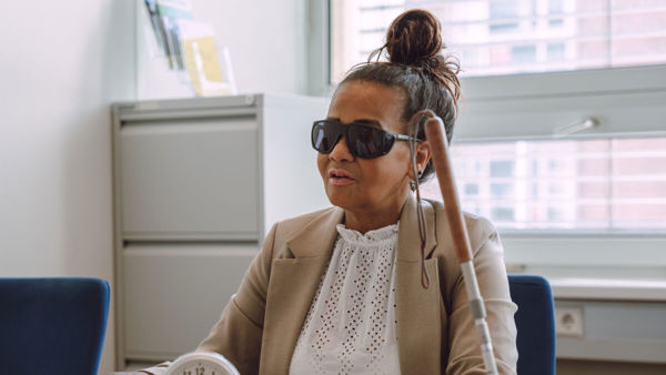 Een vrouw zit aan een tafel, in gesprek met iemand. Ze draagt een donkere zonnebril. Een taststok leunt tegen de tafel aan. Een vrouw zit aan een tafel, in gesprek met iemand. Ze draagt een donkere zonnebril. Een taststok leunt tegen de tafel aan.