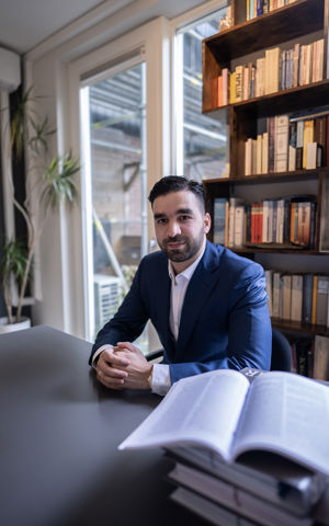 Foto van Faruk, hij zit aan een bureau in een kantoor met een blauw pak aan. Foto van Faruk, hij zit aan een bureau in een kantoor met een blauw pak aan.